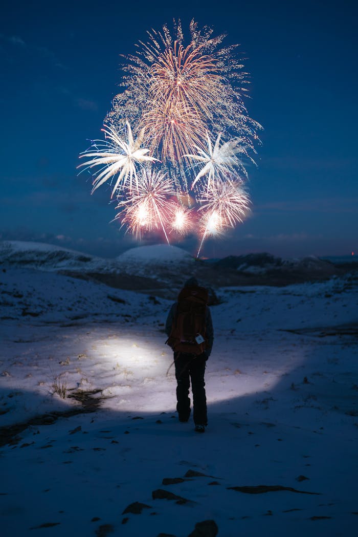 A lone hiker watches fireworks illuminating a snowy winter night, creating a magical outdoor scene.